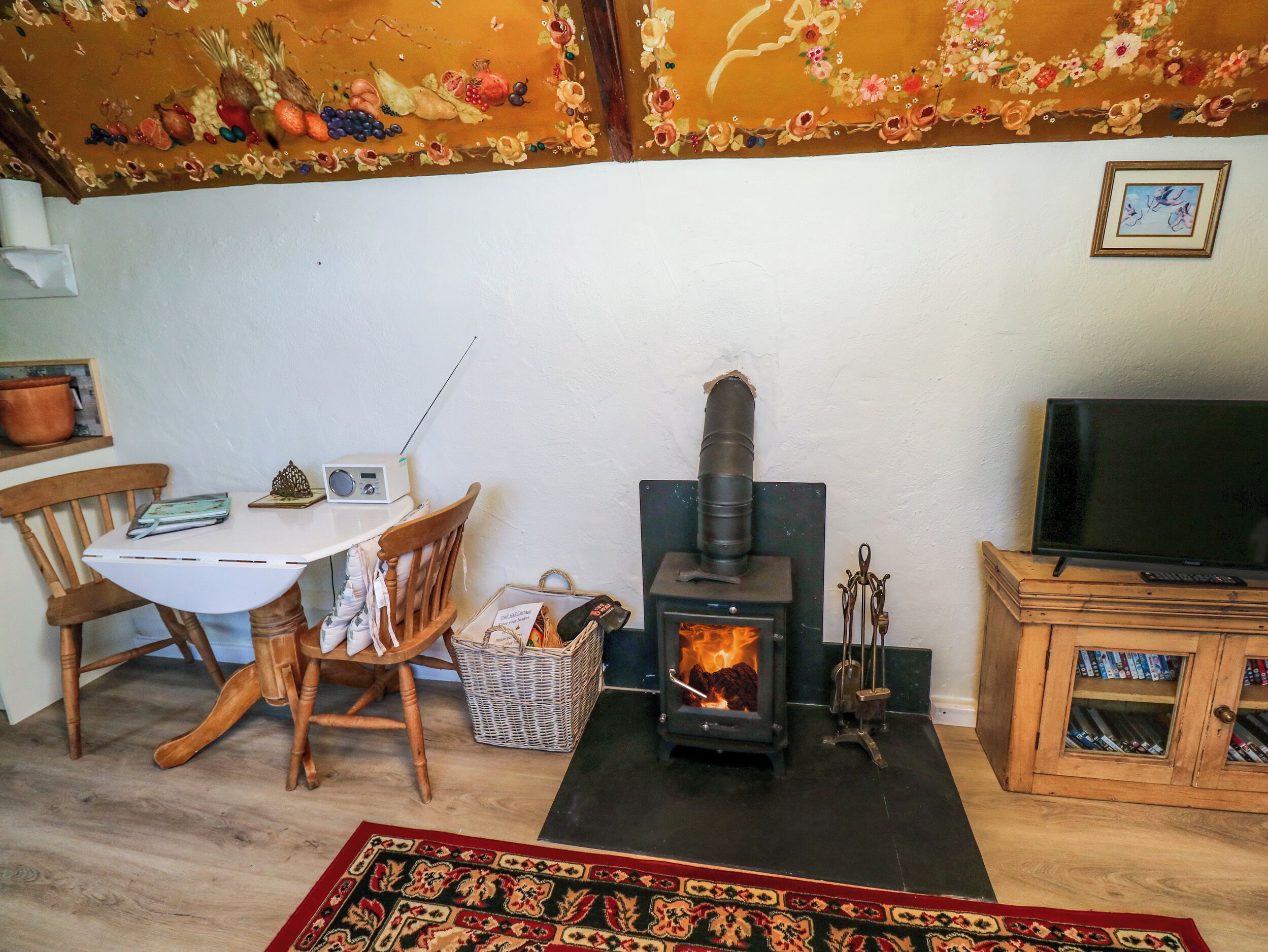 A cozy living room with a wood-burning stove, a television, a table with a chair, and a bookshelf.