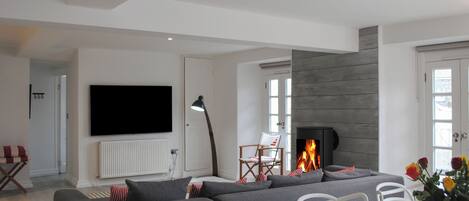 A modern living room with a large grey sectional sofa, a flat-screen TV mounted on the wall, a fireplace, and a dining area with a white table and chairs.