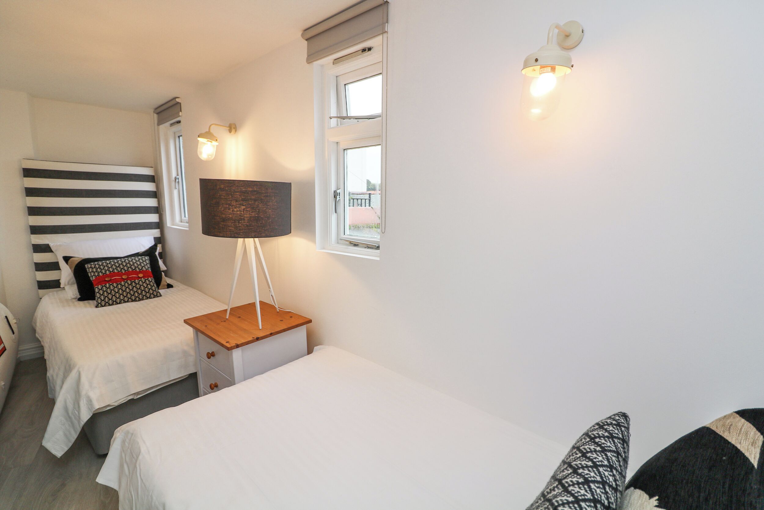 Un dormitorio con dos camas individuales, una mesita de noche con lámpara, una ventana con vistas y una lámpara de pared.