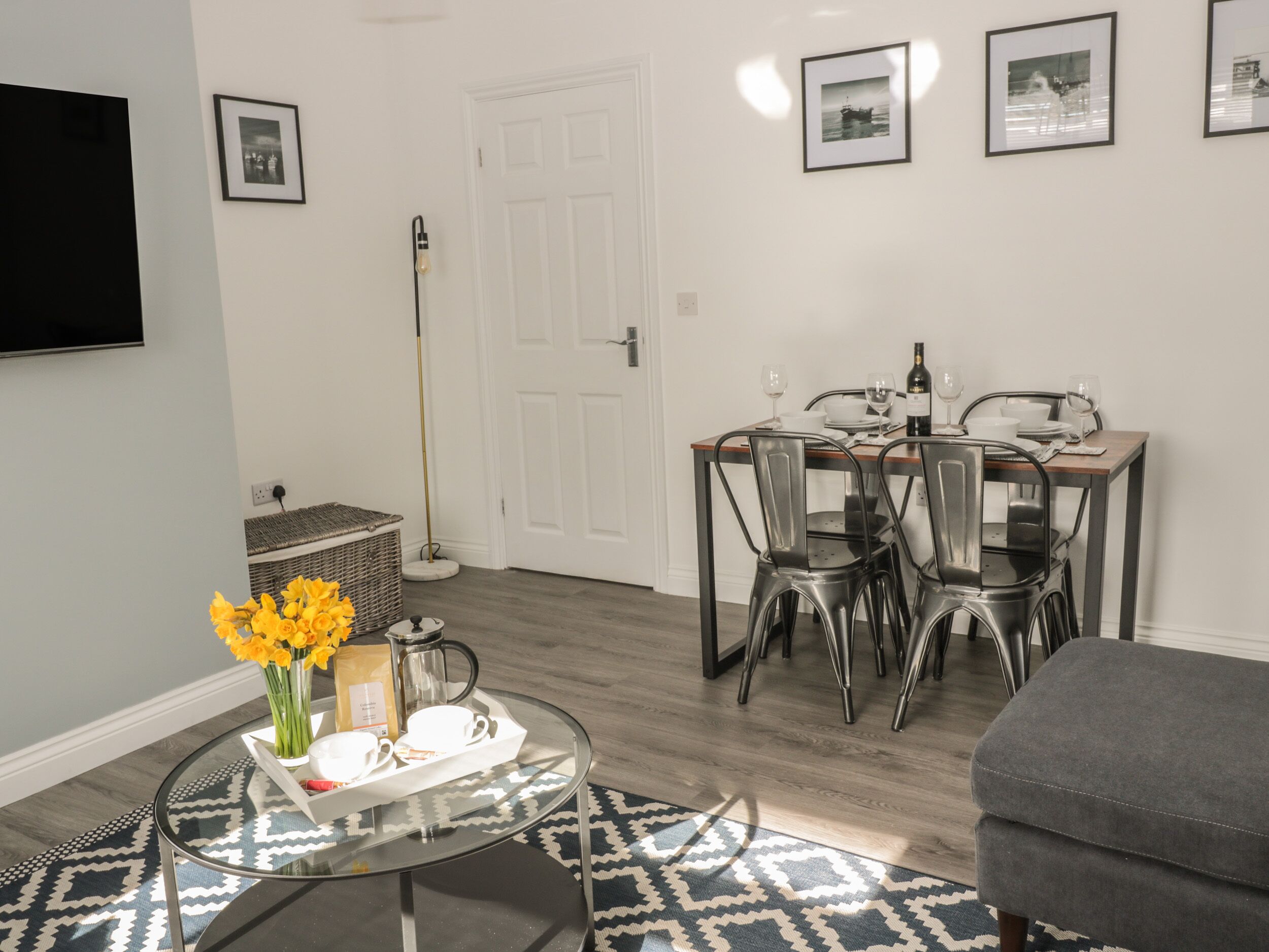 A dining area with a table set for two, chairs, a coffee pot, and a vase with yellow flowers.