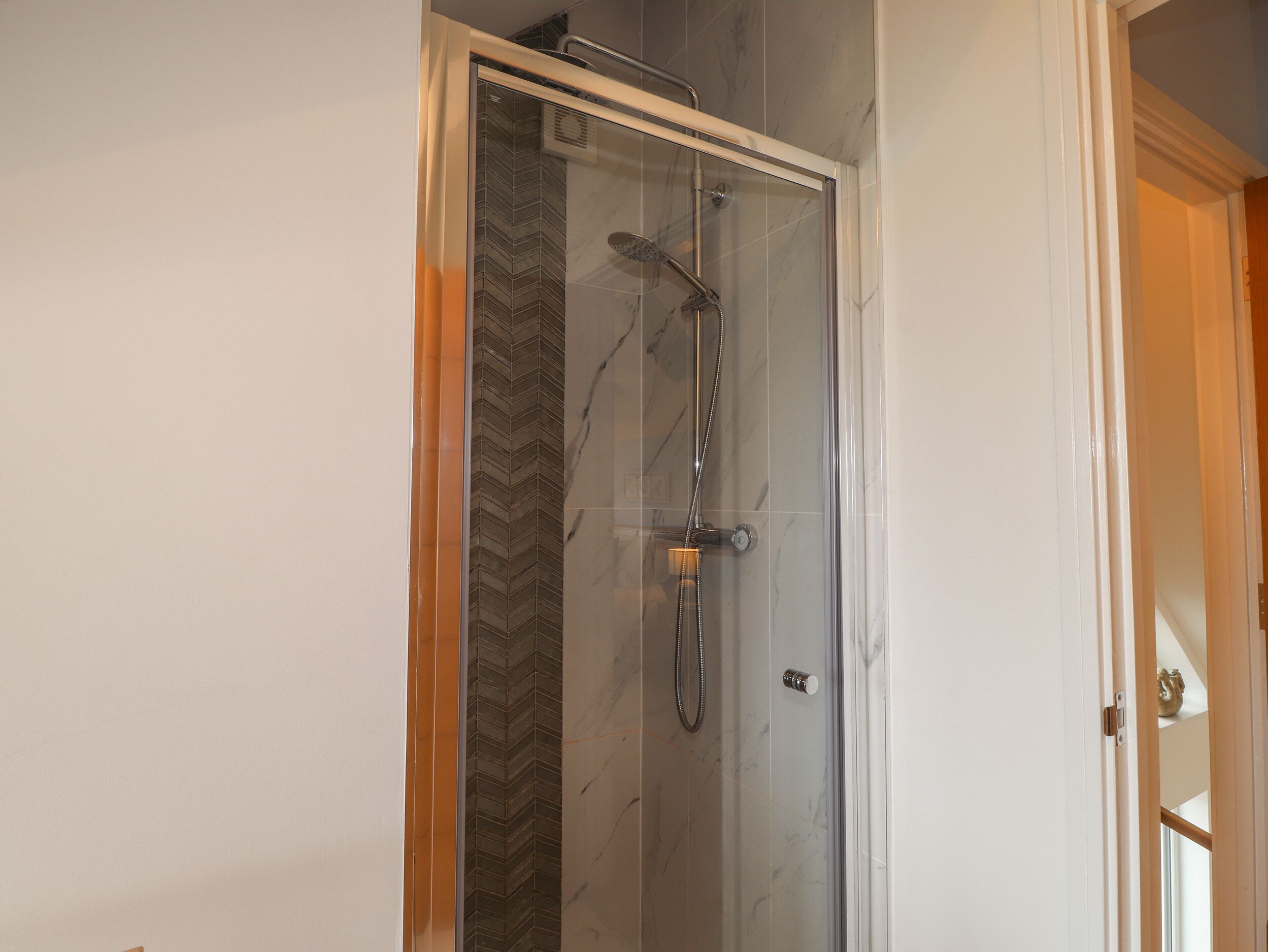 A glass-enclosed shower with a herringbone tile pattern on the wall, a handheld showerhead, and a wall-mounted faucet.