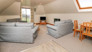 Ein geräumiges Wohnzimmer mit hoher Decke und Dachfenster, grauen Sofas, einem Holzessttisch mit Stühlen und einem Flachbildfernseher auf einem hölzernen Standfuß.