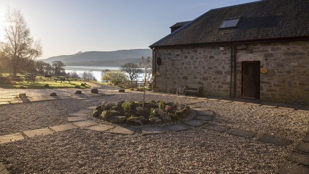 Ein Steinhaus mit Kieshof, kleinem Garten und Blick auf einen See und Berge.