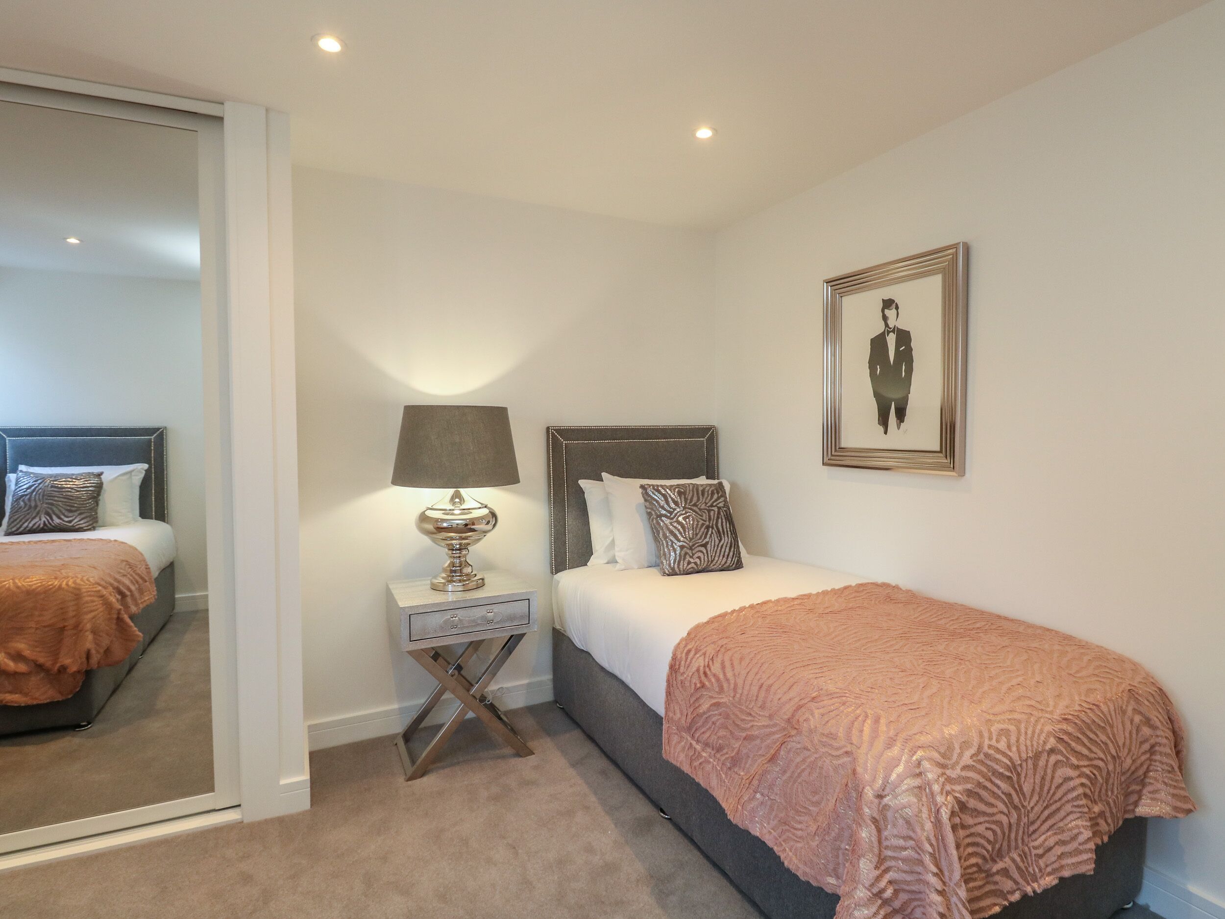 A bedroom with two beds, a bedside table with a lamp, a framed picture on the wall, and a mirror on the door.