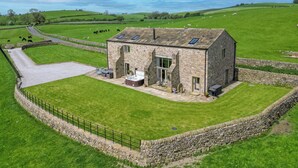 A stone farmhouse with a patio and hot tub, surrounded by a stone wall and green fields.