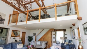 A spacious loft-style living area with a high ceiling, wooden beams, and a large glass window overlooking the interior.