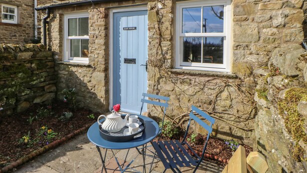 Une maison en pierre avec une porte bleue portant l’inscription « East House », un petit jardin avec une table et des chaises, et un vase rempli de fleurs.