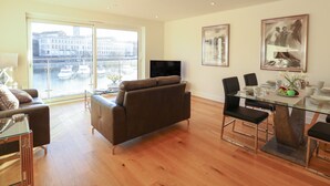 A modern living room with a large window, a brown sofa, a glass dining table, and a TV. - Harbourside Haven Apartment 4 (Weymouth)