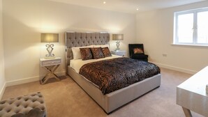 A bedroom with a tufted headboard, a bed with patterned bedding, two bedside tables with lamps, a chair, and a window. - Harbourside Haven Apartment 4 (Weymouth)