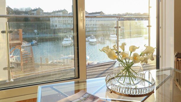 A balcony with a glass railing overlooking a canal with boats, a vase with flowers on a table, and a glimpse of buildings in the background. - Harbourside Haven Apartment 4 (Weymouth)