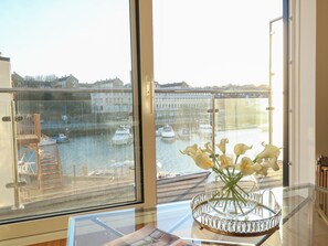 A balcony with a glass railing overlooking a canal with boats, a vase with flowers on a table, and a glimpse of buildings in the background. - Harbourside Haven Apartment 4 (Weymouth)