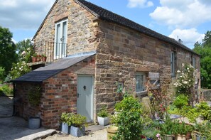 A stone house with a small balcony, a door, and a garden with various plants.