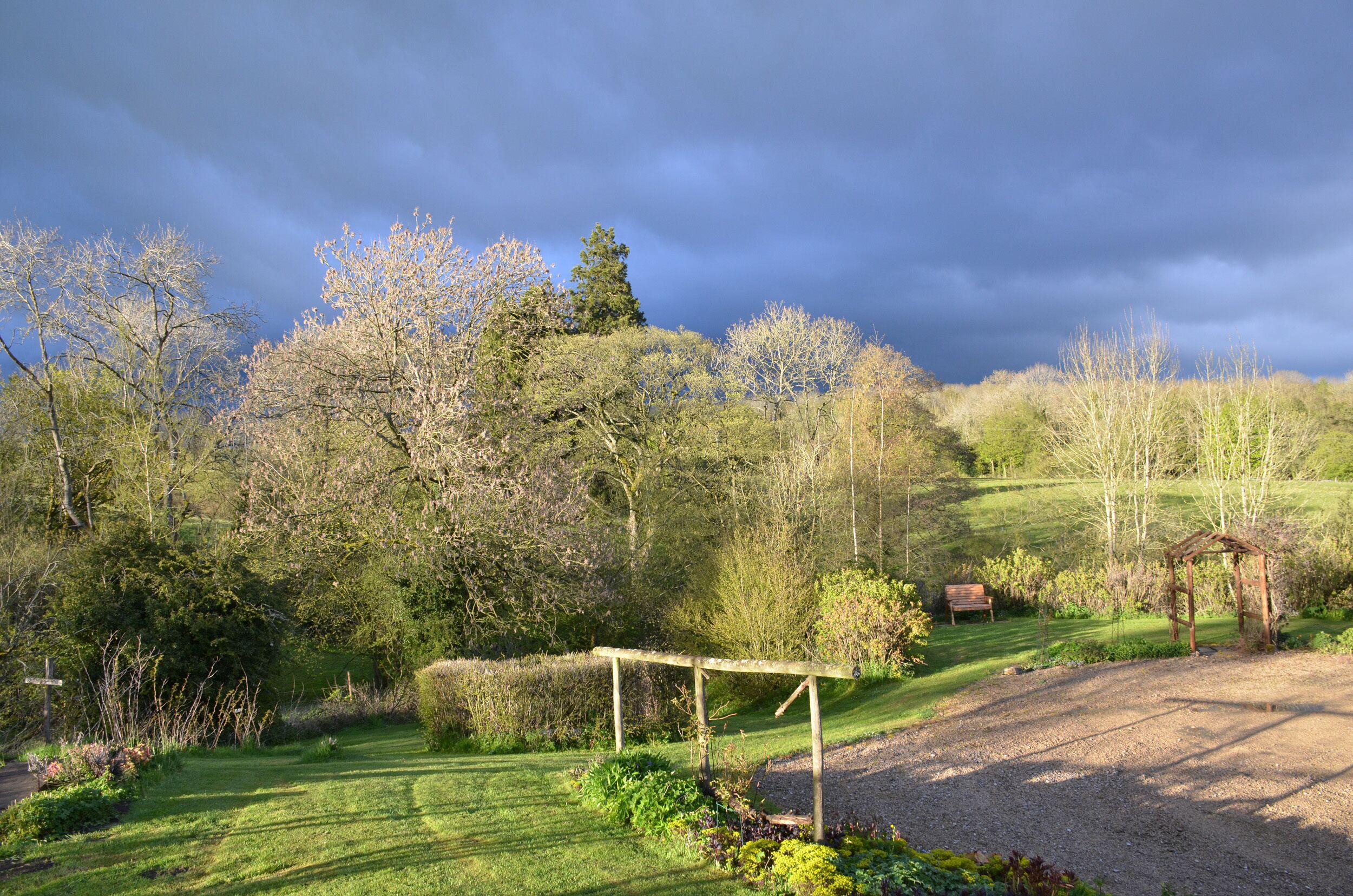 A rural landscape with a gravel path, a wooden pergola, and a bench.