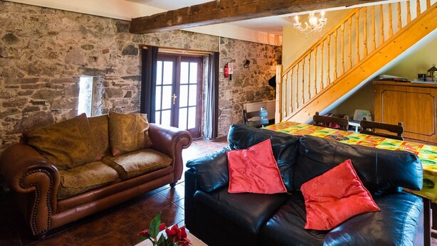 A rustic living room with a stone wall, a wooden staircase, a leather sofa, and a wooden dining table with a colorful tablecloth. - The Barn (Fethard on Sea)