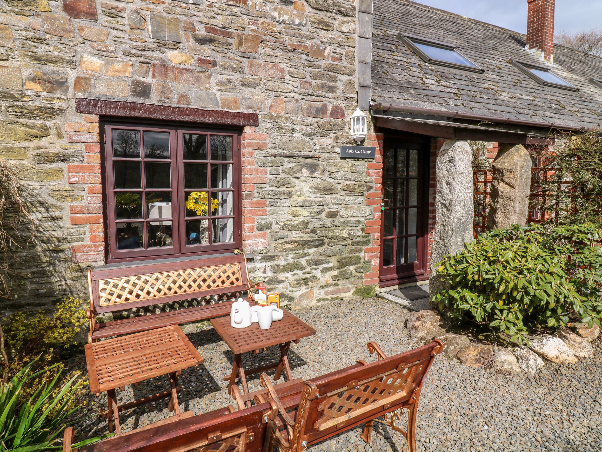 A stone cottage with a wooden bench and tables outside, a sign reading "Ash Cottage," and a small garden area.