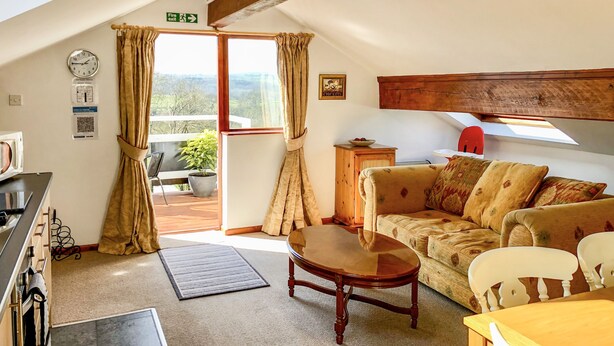 A cozy living room with a sofa, a coffee table, and a dining table. There is a window with curtains, a clock, and a fire exit sign. - Heather (Ruthin)