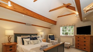 A bedroom with a large bed, wooden headboard, two bedside tables with lamps, a chair, a dresser, a television, and a window with a view of greenery. - The Coach House Apartment (Tewkesbury)