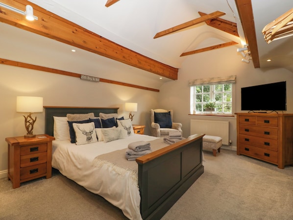 A bedroom with a large bed, wooden headboard, two bedside tables with lamps, a chair, a dresser, a television, and a window with a view of greenery.