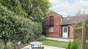 A brick house with a small balcony, a front door, and a garden area with a patio table and chairs. - The Coach House Apartment (Tewkesbury)