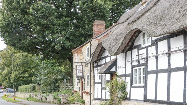 A traditional half-timbered building with a thatched roof, stone walls, and a signboard advertising ales. - The Coach House Apartment (Tewkesbury)