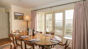 A dining room with a round table set for six, six chairs, a painting on the wall, a lamp, and a view through large windows. - Samstone (Shaftesbury)