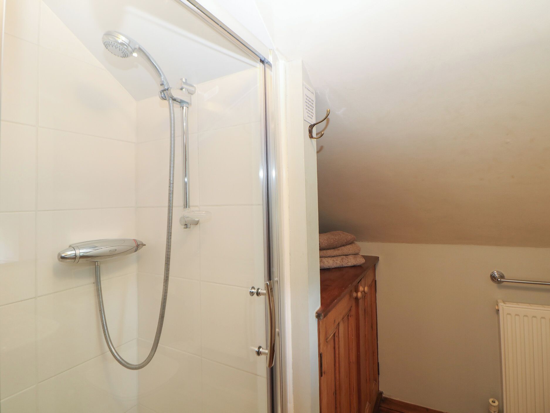 A shower with a handheld showerhead and a wall-mounted faucet, a wooden cabinet with towels, and a radiator on the wall.