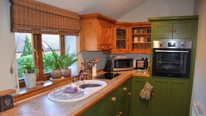 A kitchen with wooden cabinets, a microwave, an oven, a sink, and a window with plants. - Green Farm Cottage (Ashbourne)