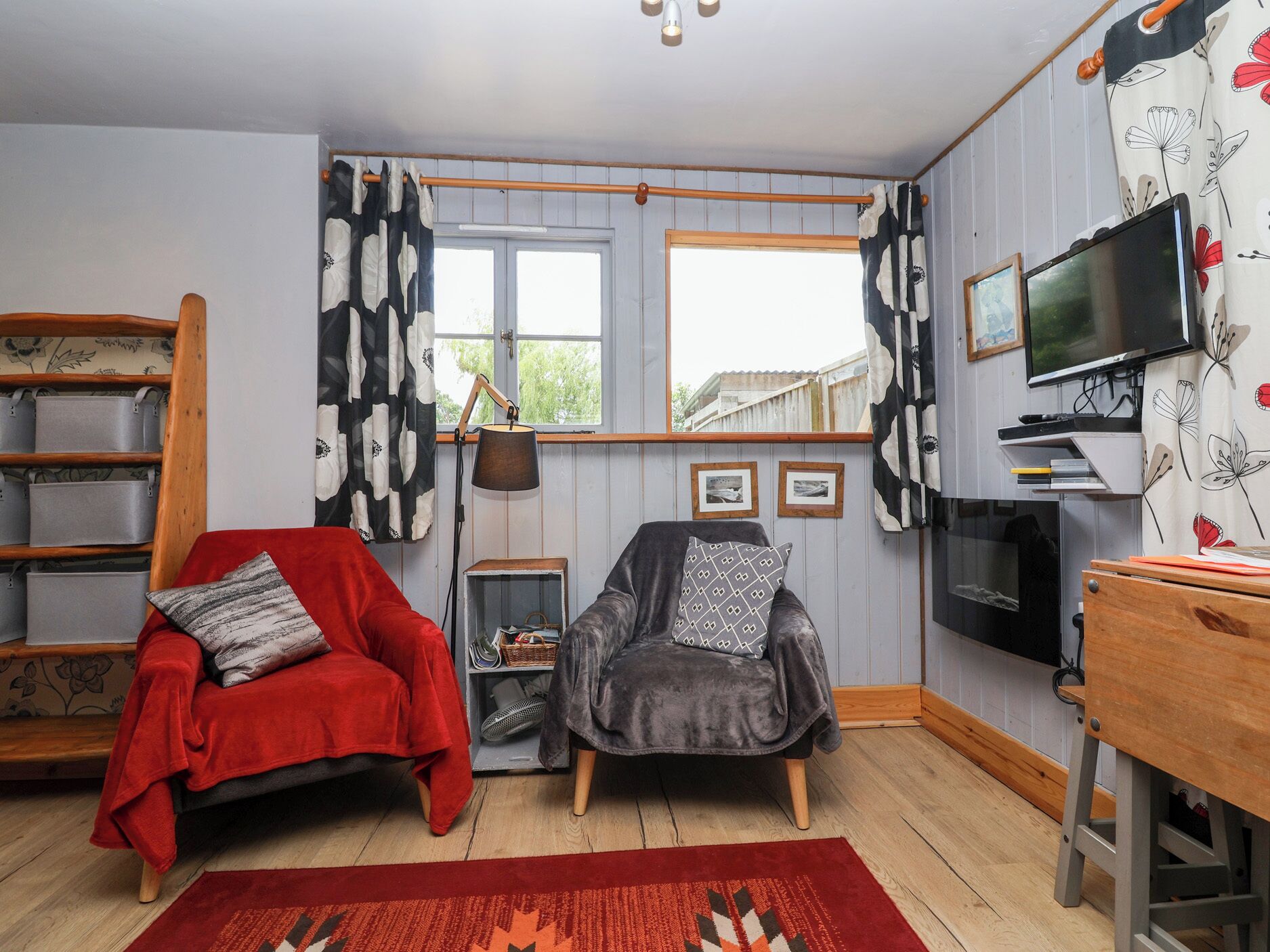 A cozy room with a red chair, a grey armchair, a wooden table, a TV, and a window with curtains.