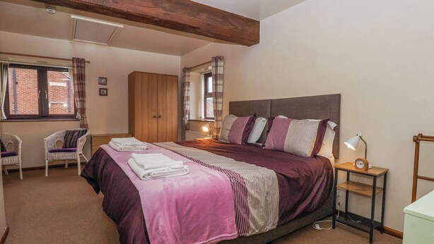 Un dormitorio con una cama grande, un armario de madera, una ventana con cortinas, una silla y una mesita de noche con lámpara y reloj.