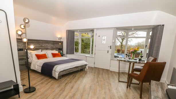 A modern bedroom with a wooden floor, a bed with white bedding and orange pillows, a bedside table, a desk with a chair, and a large window with a view of the outside. - Hockerley Cottage (High Peak)