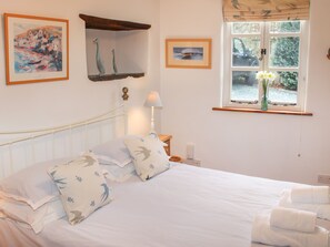 A neatly made bed with pillows, a bedside lamp, and a window with a view of greenery. - Plosh House (Keswick)
