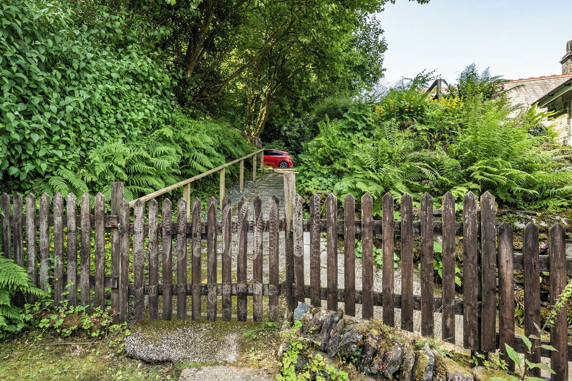 A wooden fence with a metal gate, surrounded by lush greenery and a red car parked on a stone pathway.