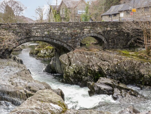 A stone bridge over a flowing river with rocky banks, surrounded by trees and buildings.