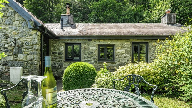 A stone house with a chimney, a window, and a well-maintained garden with a round metal table and wine bottles. - Tyn Y Ffynnon (Betws-Y-Coed)