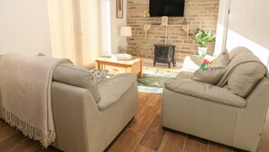 Un salon avec deux canapés beige, une table basse en bois, une cheminée surmontée d’un téléviseur et un mur en briques.