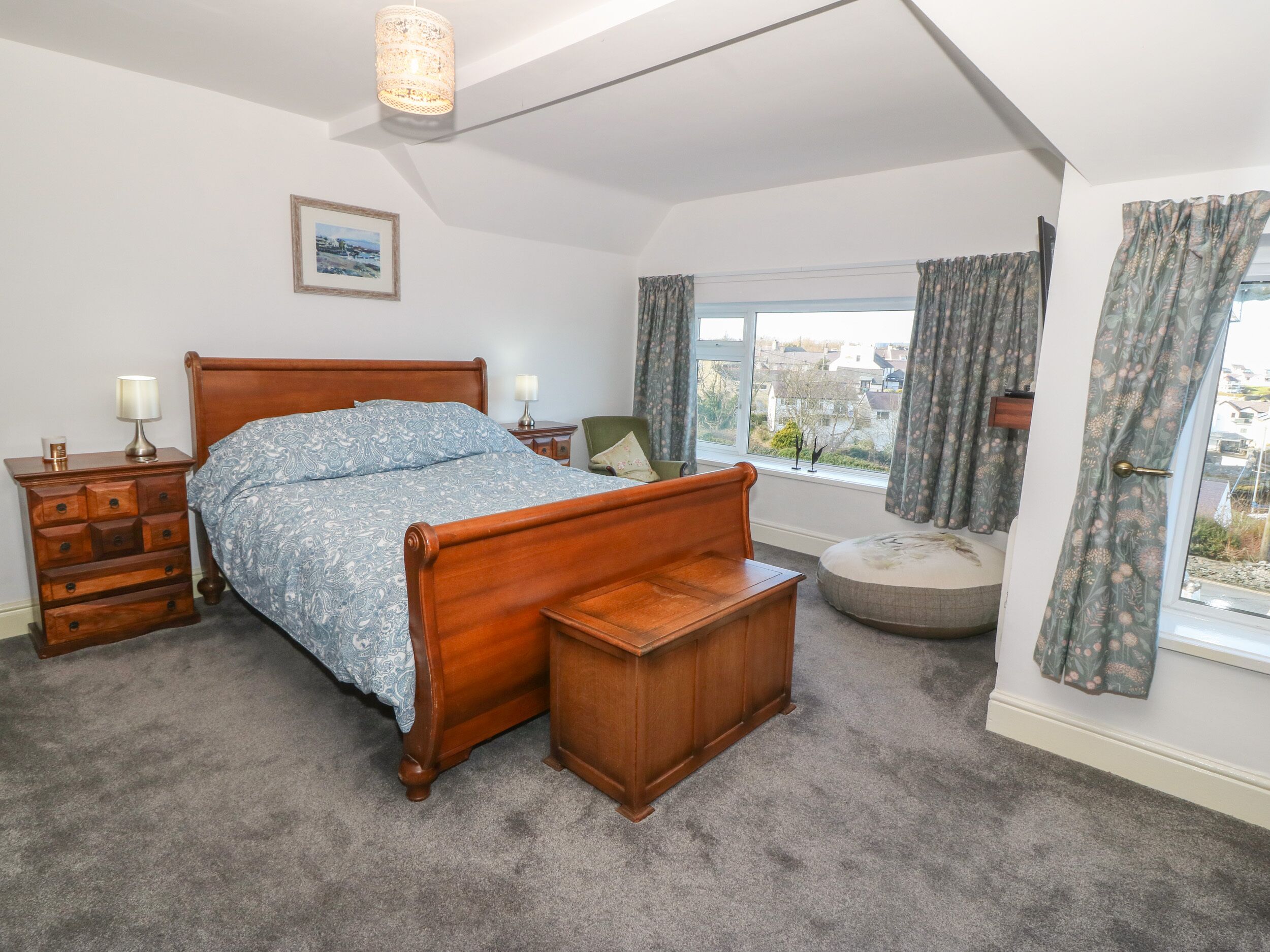 A bedroom with a wooden bed, a nightstand with a lamp, a dresser, a window with curtains, and a view of buildings outside.