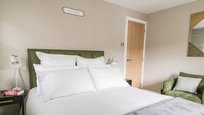 A neatly made bed with white linens and a grey blanket, a green upholstered chair, a bedside table with a lamp, and a door with a wooden panel. - 126 Main Street (Keighley)