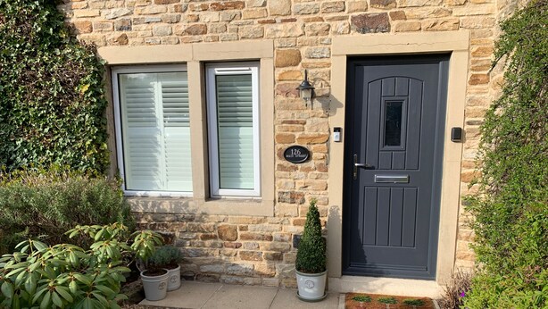 A stone house with a black door, a window with shutters, and a small potted plant beside the door. - 126 Main Street (Keighley)