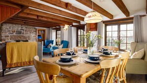 A cozy dining area with a wooden table set for four, surrounded by chairs. A stone wall and wooden beams are visible in the background. - Little Leighs (Launceston)