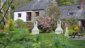 A traditional stone house with a white exterior, surrounded by a well-maintained garden with flowering plants and a stone bench. - Little Leighs (Launceston)