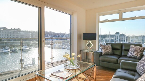 Un salon spacieux avec une table basse en verre, un canapé en cuir et une vue sur une marina, grâce à de grandes fenêtres.