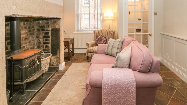 A cozy living room with a fireplace, a sofa, a lamp, and a window with blinds. - Chapel Cottage (Corbridge)