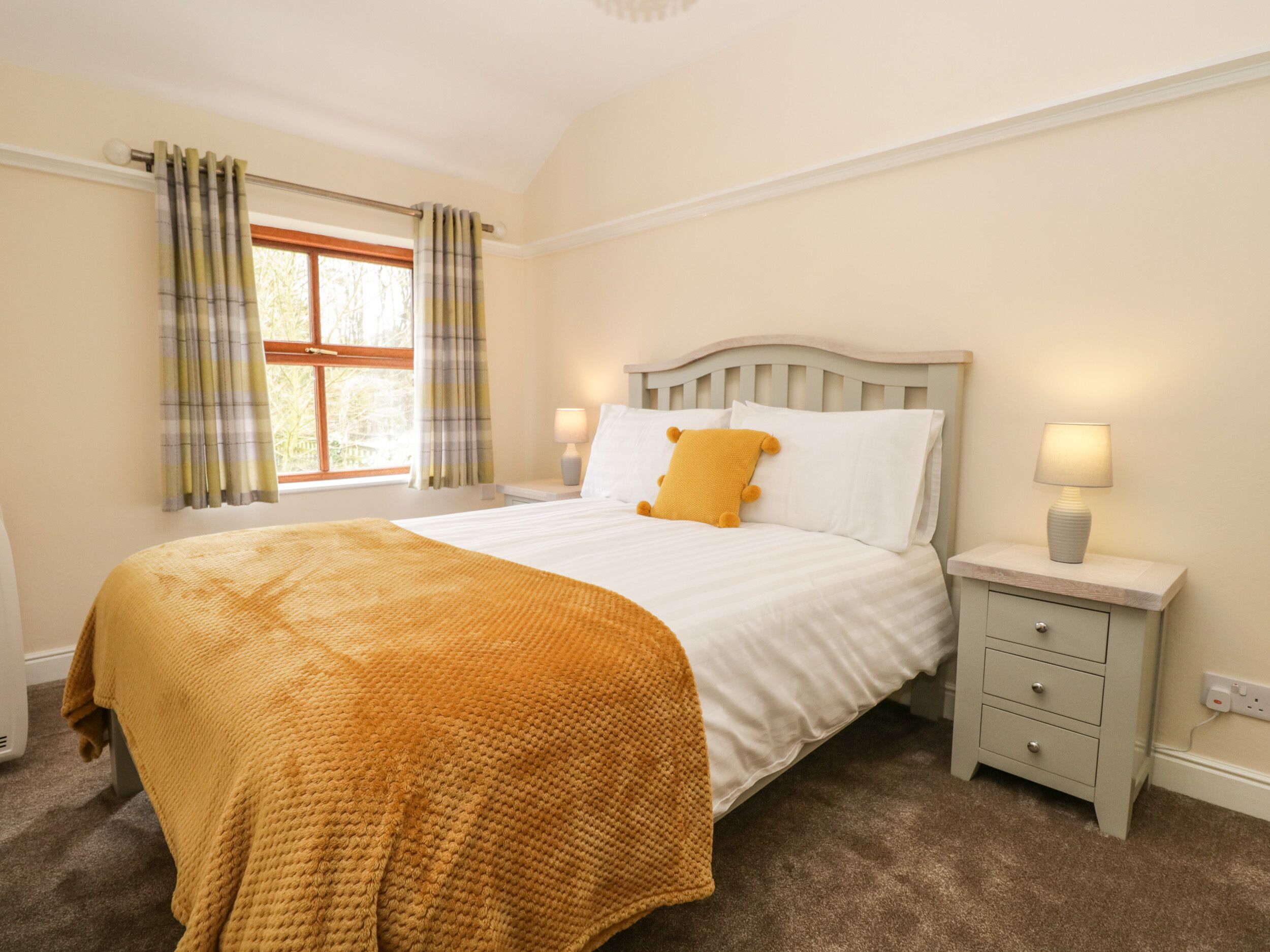 A bedroom with a bed, a bedside table, a lamp, a window with curtains, and a carpeted floor.