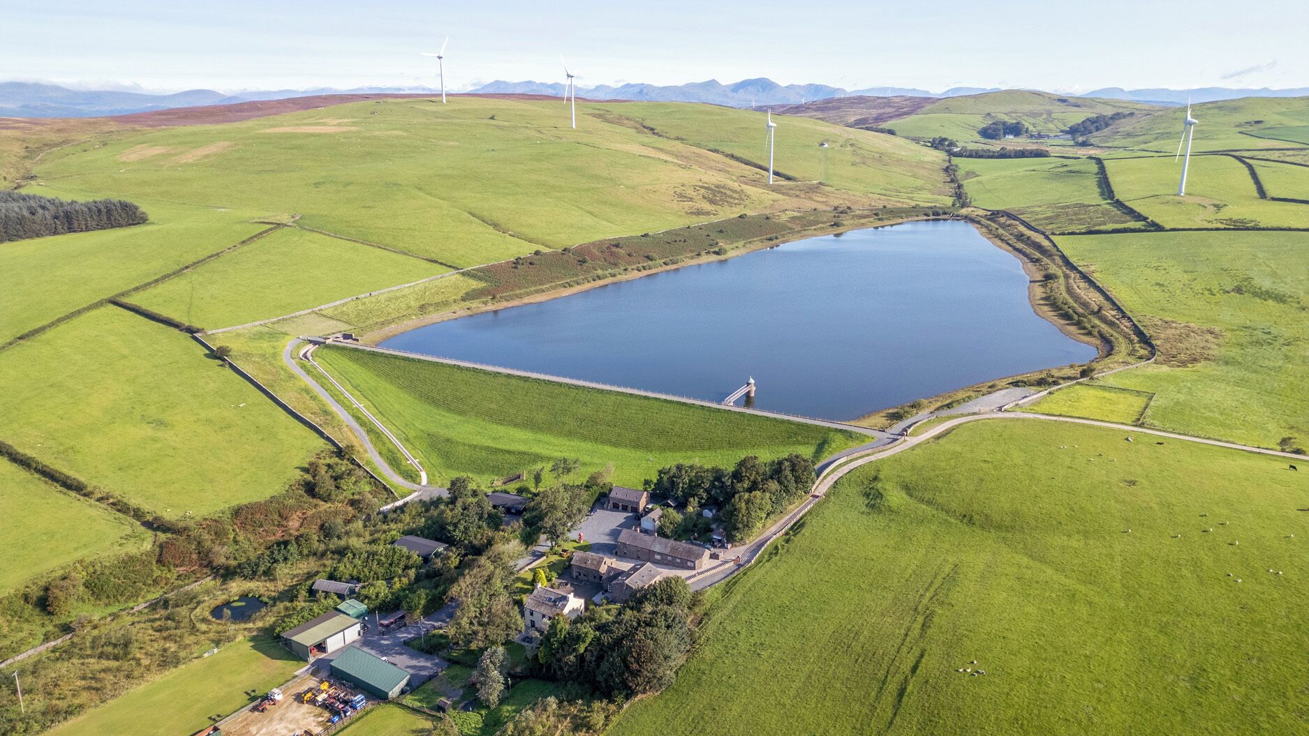 Vista aérea de uma área rural com um lago grande, turbinas eólicas e um pequeno povoado com edifícios e uma estrada.