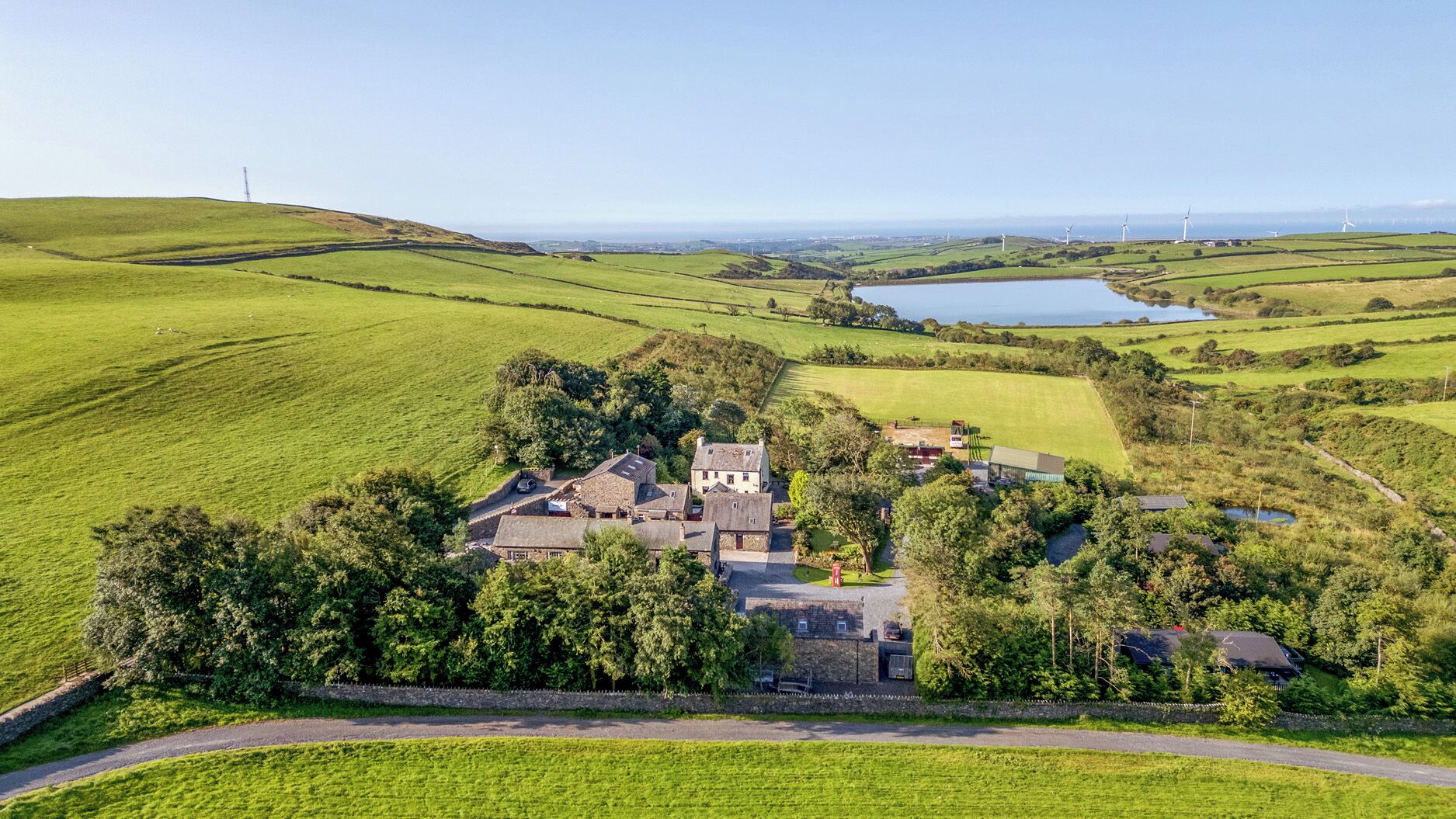 A rural property with multiple buildings, a pond, and wind turbines in the background.