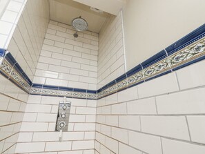 Esquina de un baño con azulejos blancos, un cabezal de ducha y detalles decorativos con azulejos azules y verdes a los bordes.