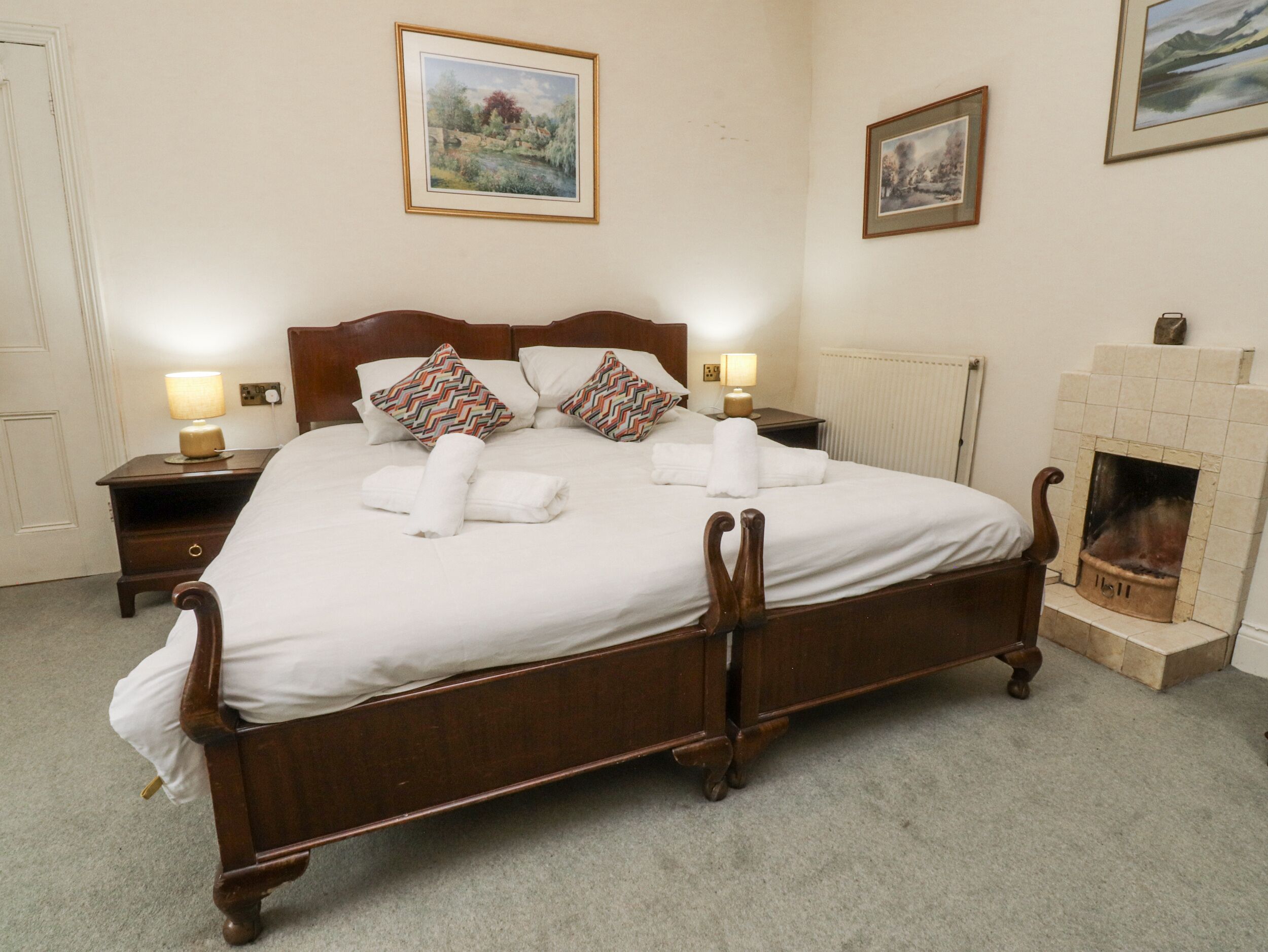 Uma cama bem arrumada com lençóis brancos e almofadas decorativas, cabeceira de madeira, mesas de cabeceira com abajures e quadros nas paredes.