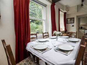 Una mesa de comedor preparada para cuatro personas, con manteles blancos, cortinas rojas y un jarrón con flores.