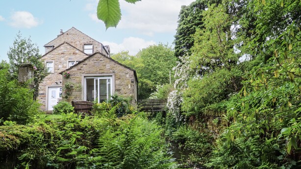 A stone house with a wooden door and windows, surrounded by lush greenery and a small stream. - Bridgend Cottage (Skipton)