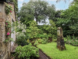 Uma trilha de pedra serpenteia por um jardim exuberante, repleto de plantas diversas e uma construção rústica em pedra.